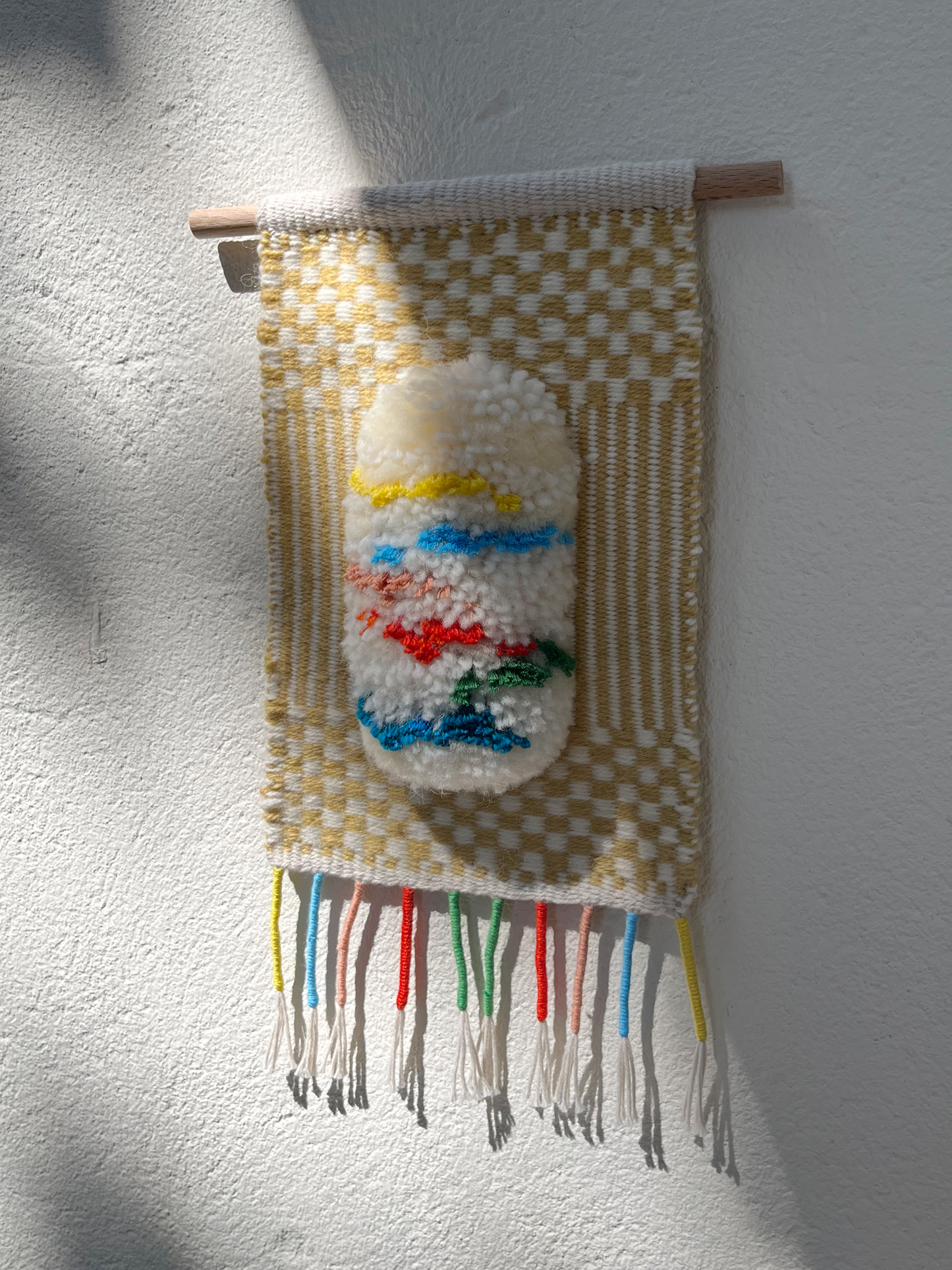 A handmade woven wall hanging with a cream and tan checkerboard pattern, featuring a central raised white shape with blue, red, green, and yellow accents, suspended from a wooden dowel against a white textured wall.