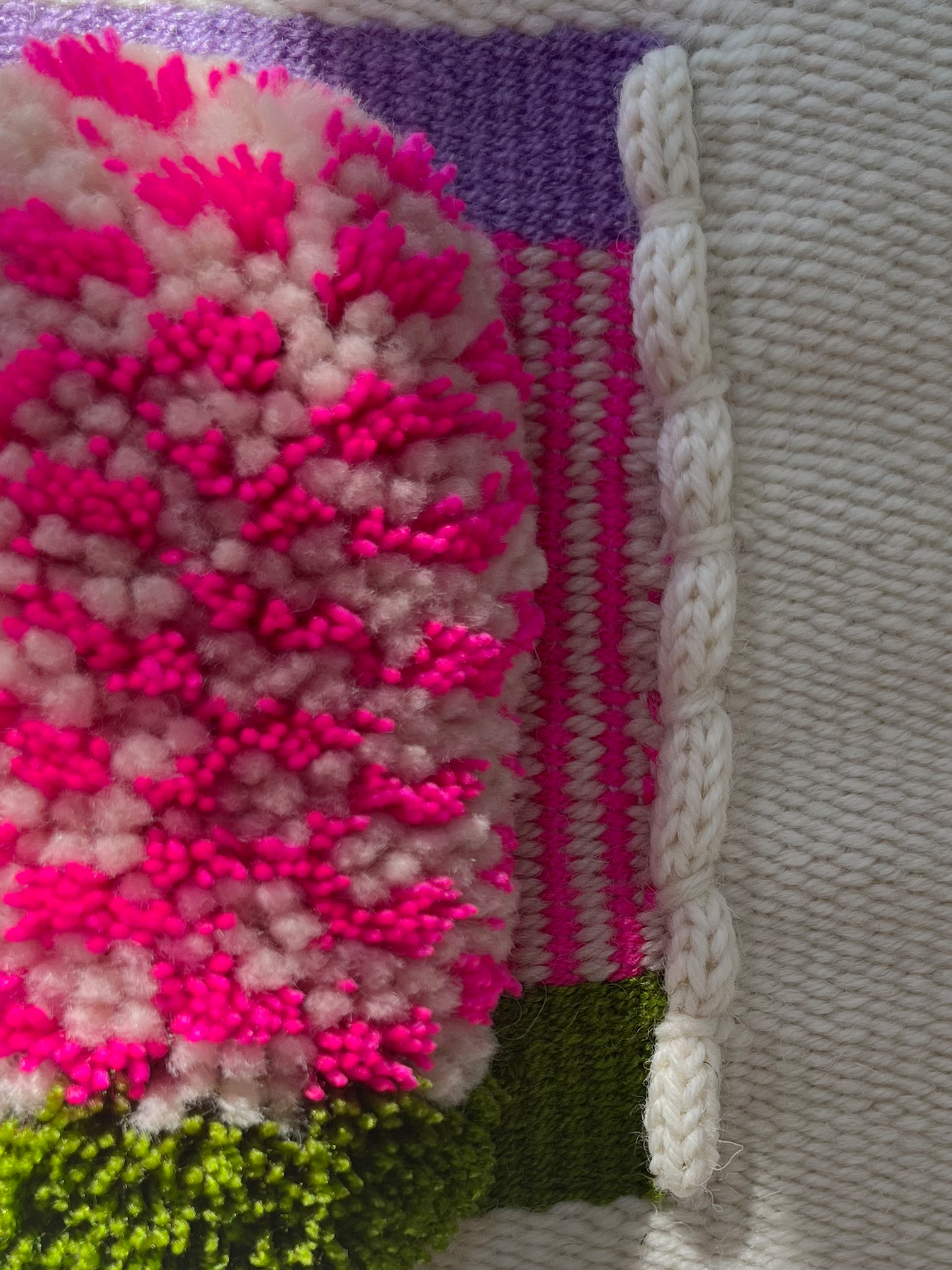 Close-up of a colorful handwoven textile featuring a large fluffy pom-pom texture in pink, white, and magenta, alongside knitted sections in purple, white, green, and bright pink.