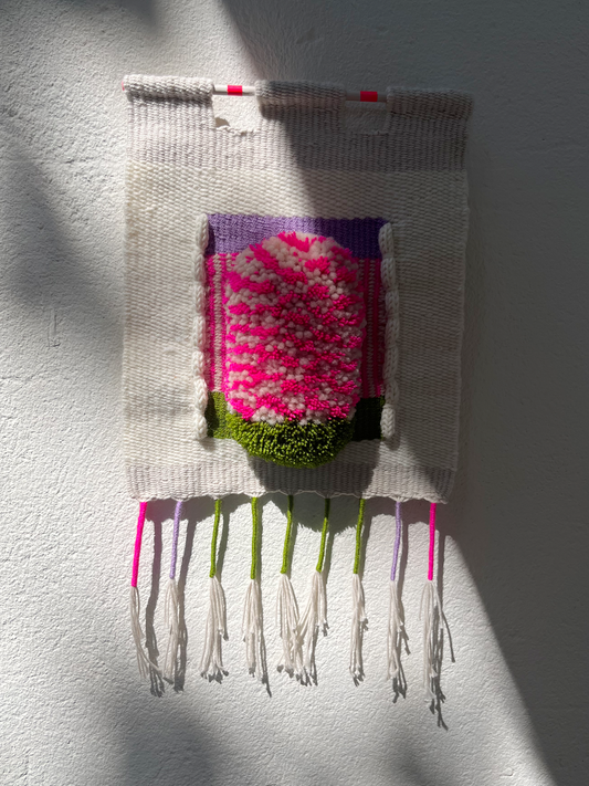 A handwoven textile wall hanging with a central floral motif in bright pink, white, purple, and green, suspended against a white wall with dramatic natural shadows.