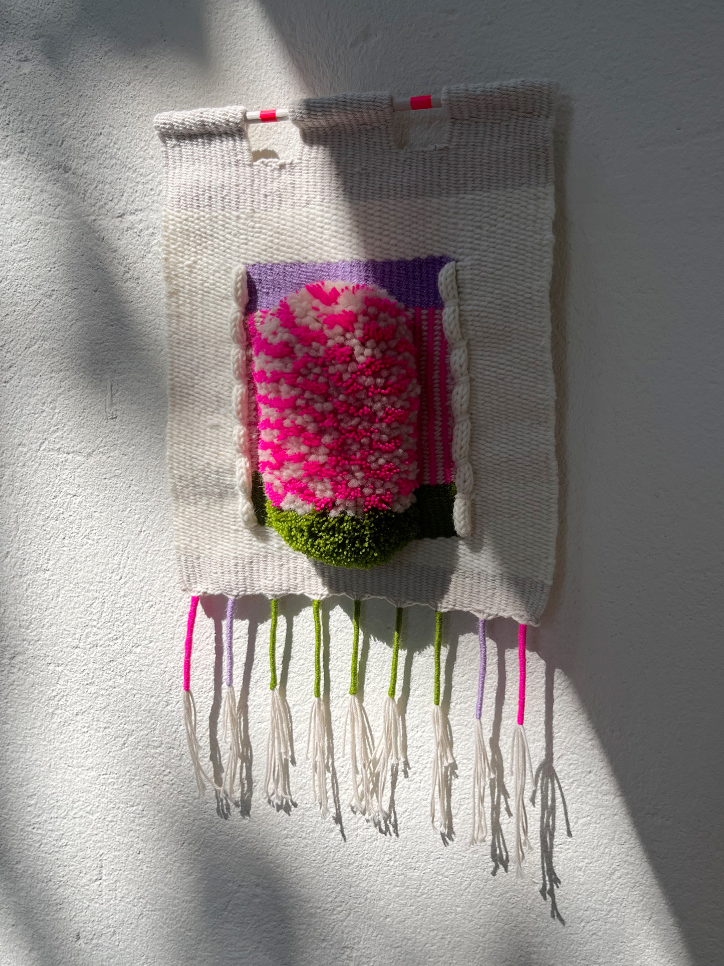 A handwoven wall hanging textile with a central raised floral motif in bright pink, white, purple, green, and black, suspended against a white wall with sunlight and shadows.