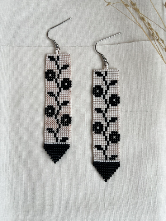 A pair of long rectangular beaded drop earrings with silver fishhook wires, featuring a black and white floral vine pattern on a cream background, finished with black triangular tips, displayed on a textured beige linen fabric.
