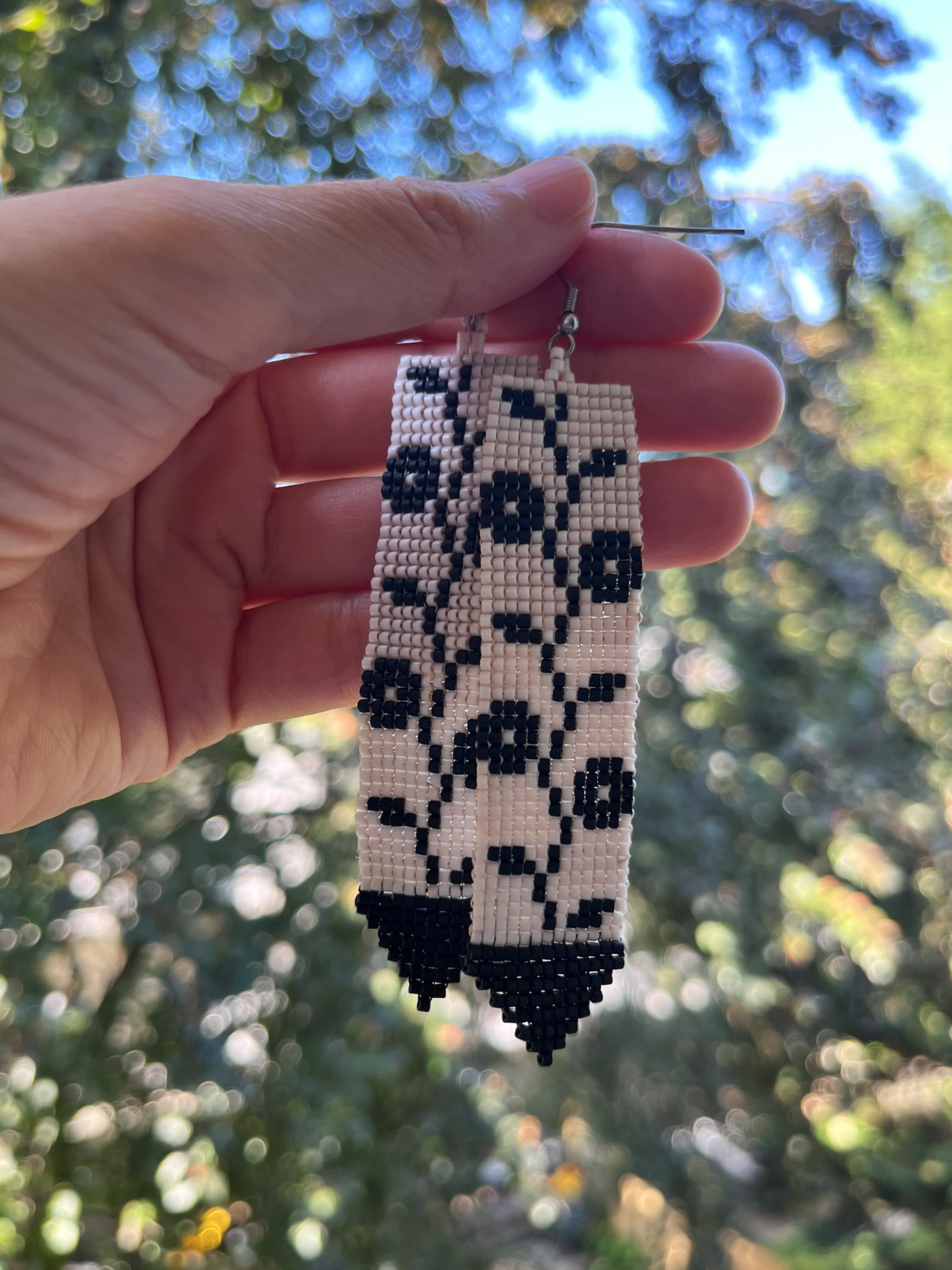 A hand holds a pair of long rectangular beaded earrings with a black floral pattern on a cream background, photographed outdoors against a blurred green and blue bokeh background.