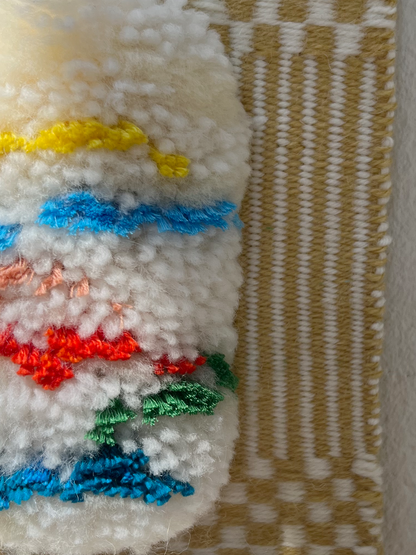 Close-up of a white textured woven hanging with colourful stripes in yellow, blue, red, green, and pink, next to a beige and white woven textile.