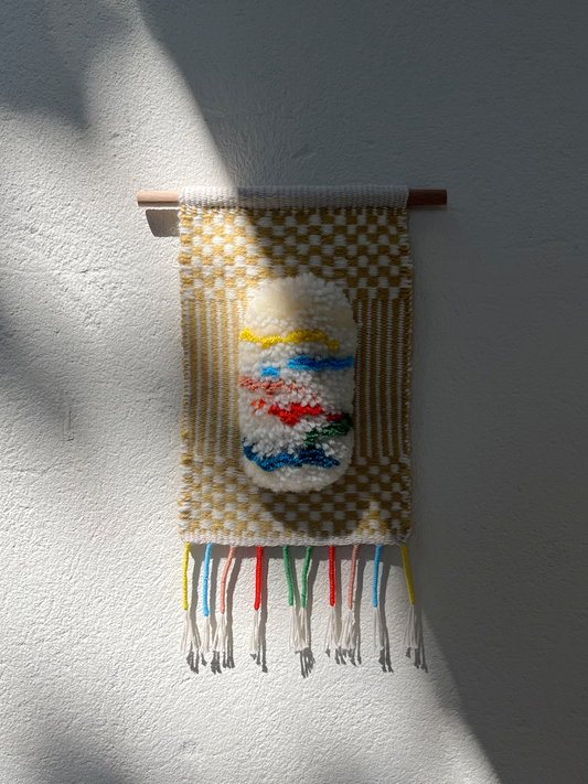 White textured woven hanging with colourful stripes in yellow, blue, red, green, and pink, next to a beige and white woven textile.