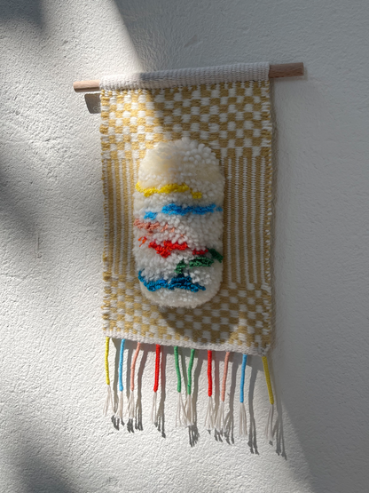 A handmade woven wall hanging with a cream and tan checkerboard pattern, featuring a central raised white shape with blue, red, green, and yellow accents, suspended from a wooden dowel against a white textured wall.