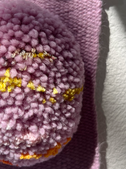 Close-up of a pink fuzzy textured weaving, with visible gold and warm thread details, next to a pink background and a white surface.