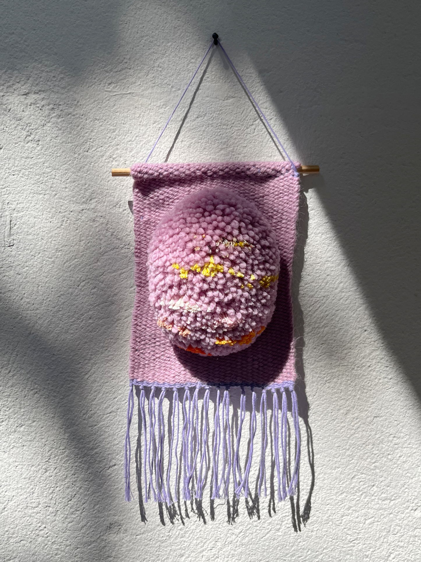 A pink woven wall hanging with fringe, suspended from a wooden dowel against a textured white wall. The center features a raised, fuzzy pink rounded element with small yellow and orange details.