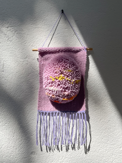 A pink woven wall hanging with fringe, suspended from a wooden dowel against a textured white wall. The center features a raised, fuzzy pink rounded element with small yellow and orange details.