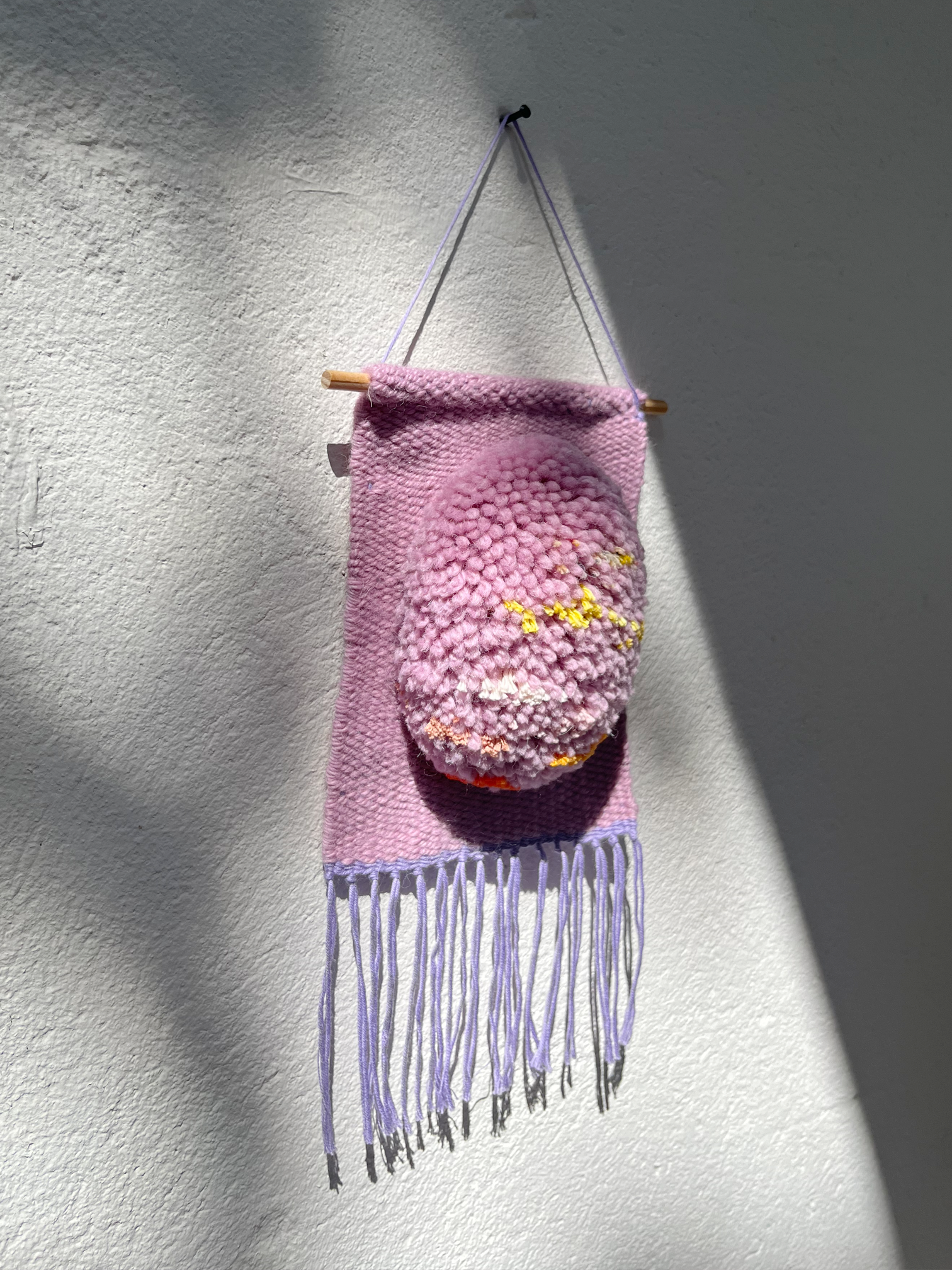A handmade lilac woven wall hanging with fringe and a small hanging oval patch featuring colorful details, displayed against a white textured wall with strong sunlight and shadows.