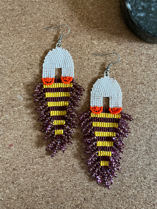 A pair of handmade beaded earrings shaped like small fish, featuring white bead tops, orange accents, yellow and white striped bodies, and dark purple dangling fringed sections, displayed on a cork surface.