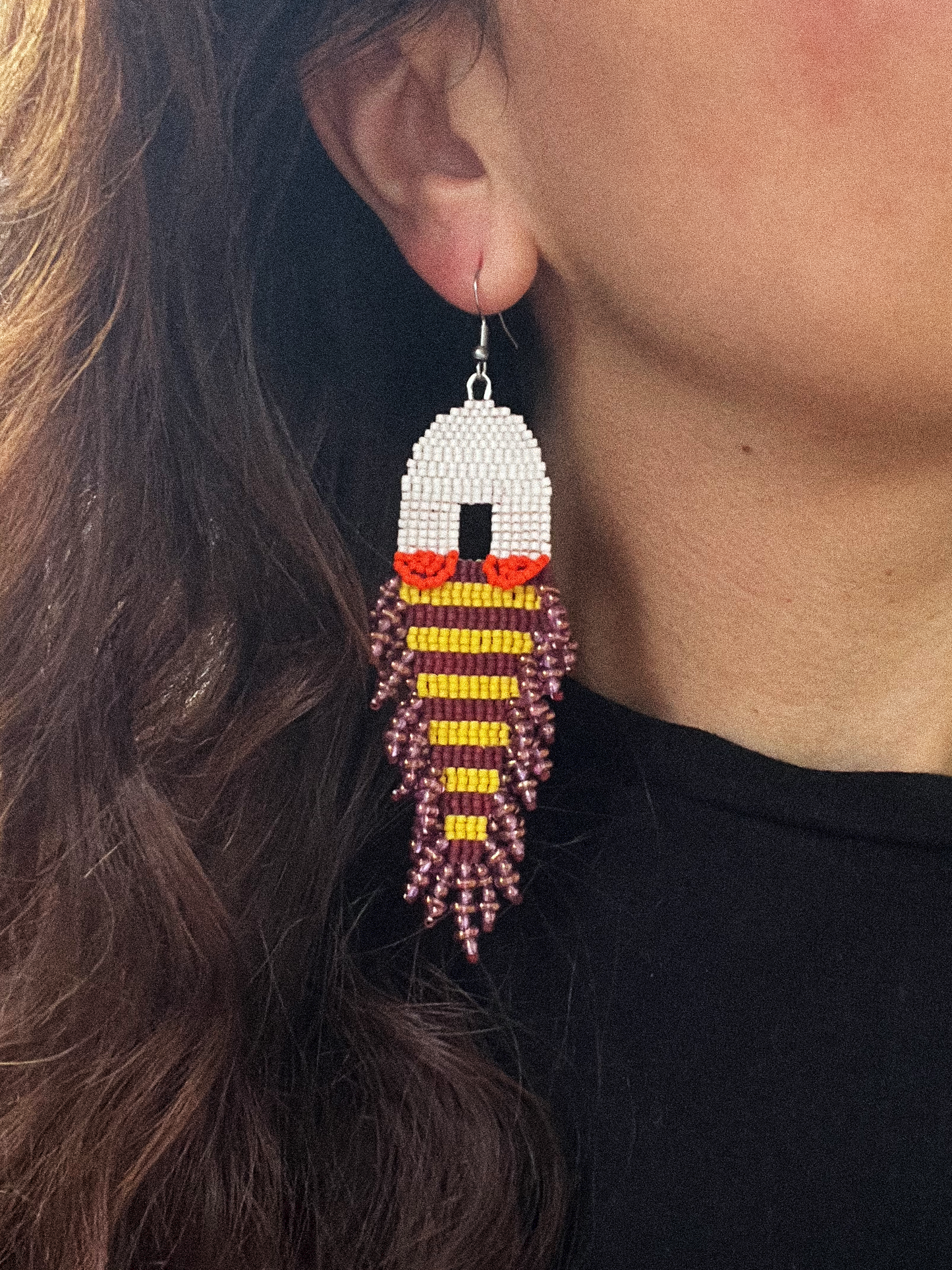 A close-up of a person wearing a long beaded dangle earring. The earring features a cream top section, followed by yellow and currant striped sections, and a dangling burgundy beaded fringe.