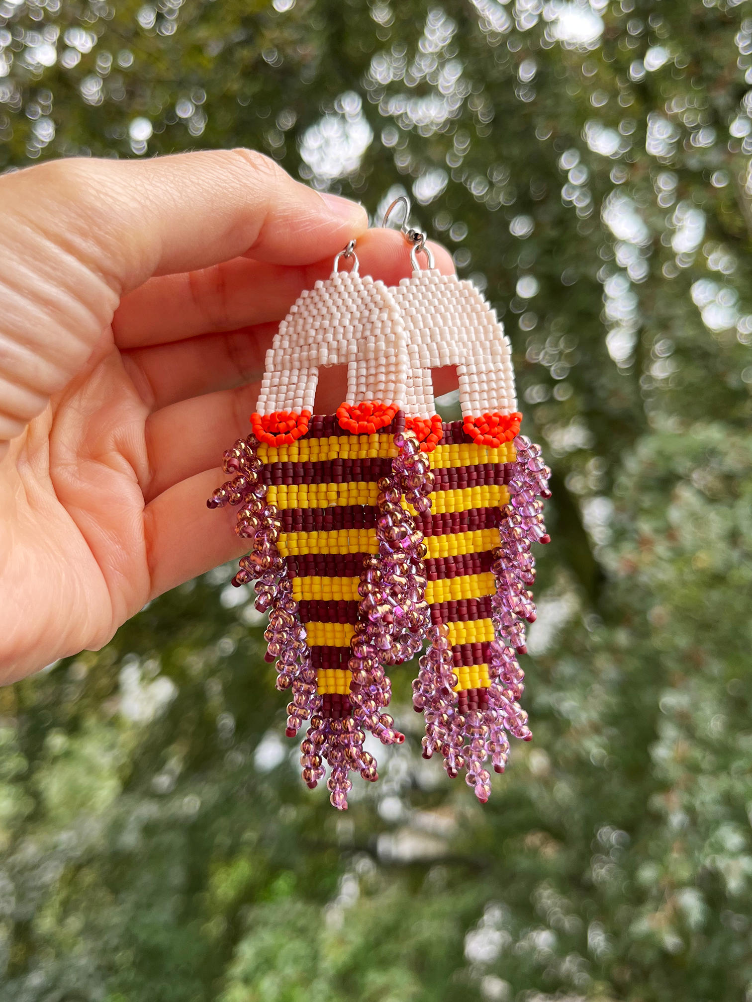 Hand holding a pair of beaded drop earrings with cream arches, orange-red accents, and yellow and dark red horizontal stripes, with dangling purple and pink beaded fringe, against a blurred green outdoor background.