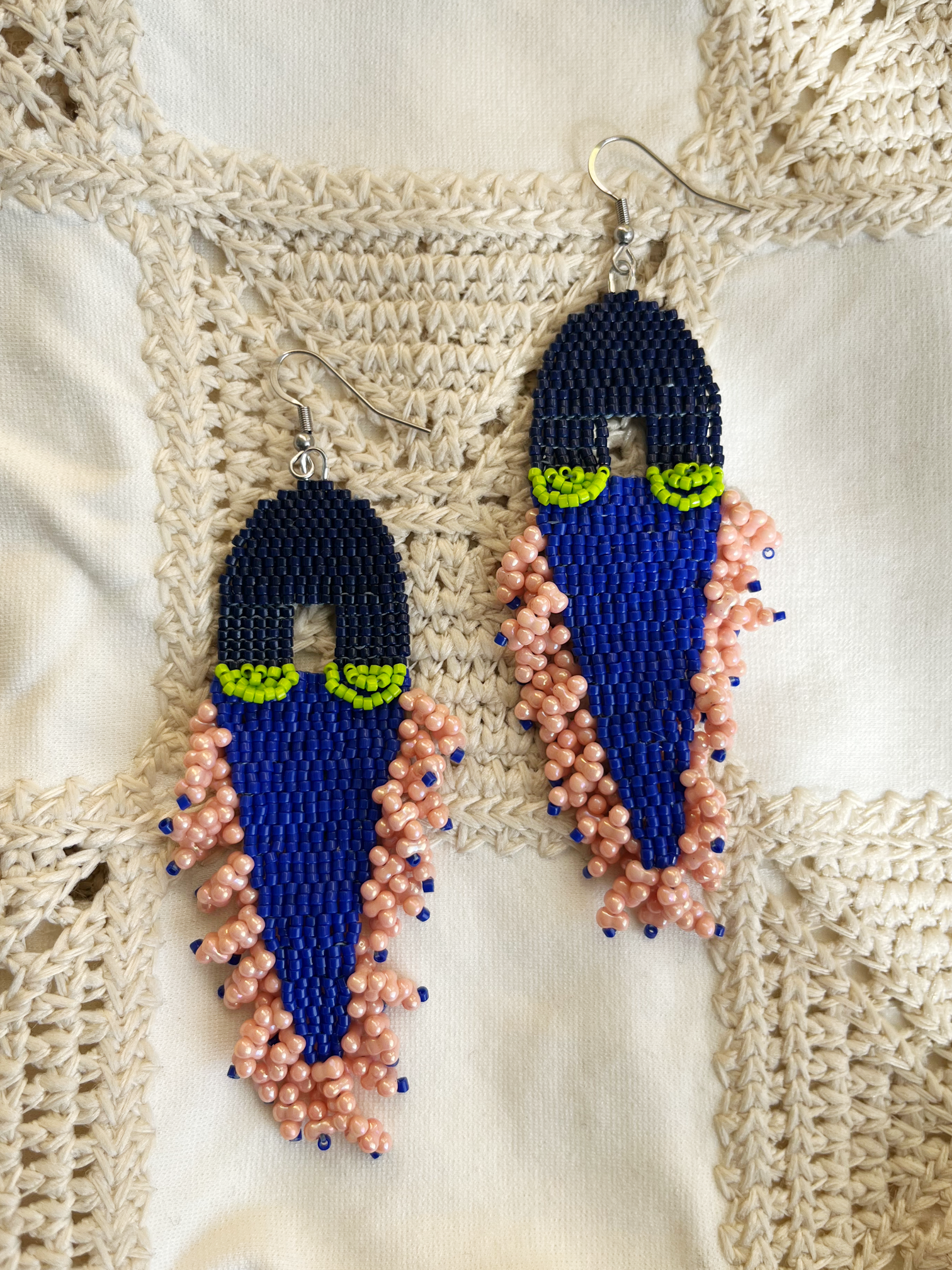 A pair of handmade beaded dangle earrings featuring a dark blue top section, a bright royal blue middle section, lime green eye-like accents, and peach-pink beaded fringe, displayed on a cream crochet background.