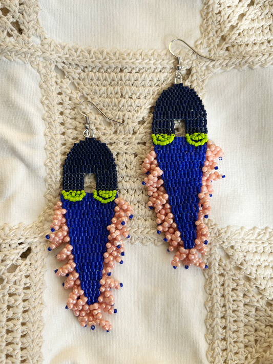 A pair of handmade beaded dangle earrings featuring a dark blue top section, a bright royal blue middle section, lime green eye-like accents, and peach-pink beaded fringe, displayed on a cream crochet background.