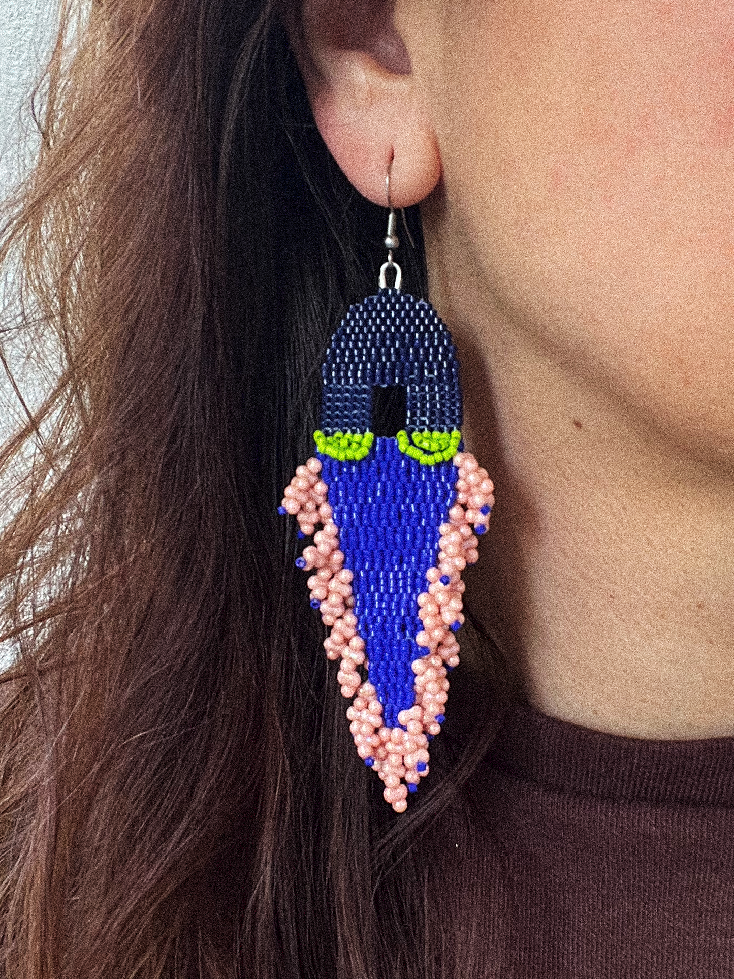 A close-up of a person wearing a large beaded dangle earring. The earring features a dark blue top section with small green accents, a bright blue middle section, and pink beaded fringe at the bottom.
