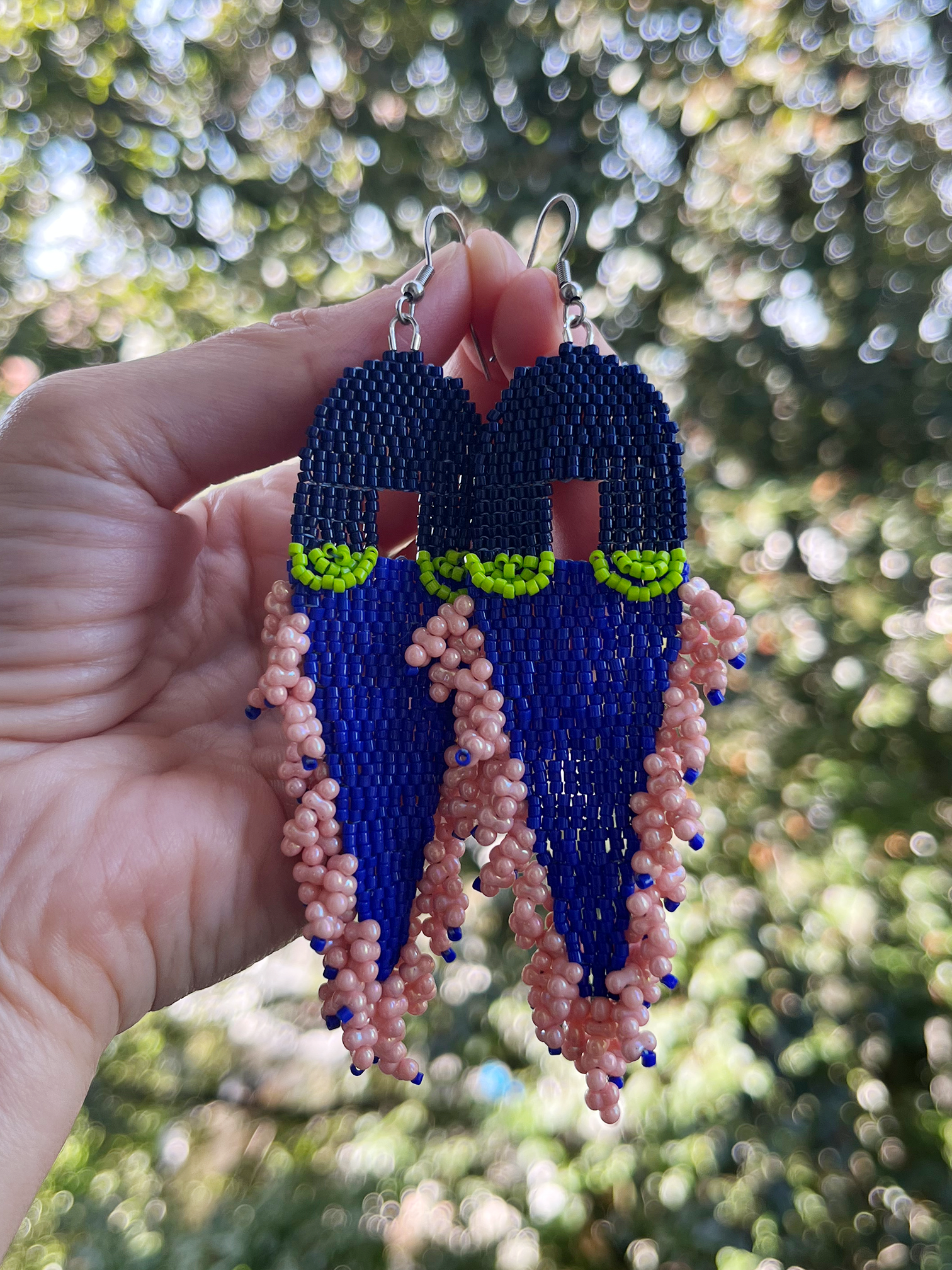 A pair of handmade beaded earrings held by a hand against a blurred outdoor background of green foliage. The earrings feature a dark blue top section, a bright lime green middle band, and a long dangling fringe made of pink and blue beads.