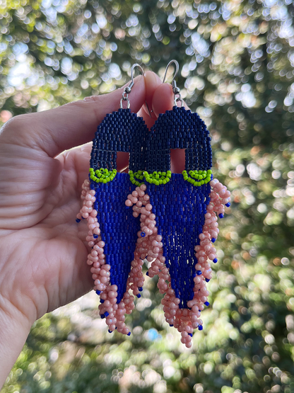 A pair of handmade beaded earrings held by a hand against a blurred outdoor background of green foliage. The earrings feature a dark blue top section, a bright lime green middle band, and a long dangling fringe made of pink and blue beads.