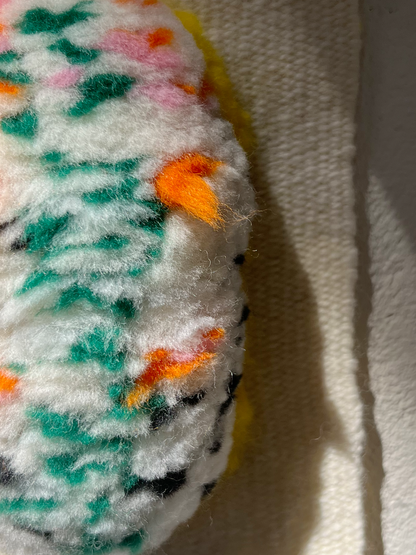 close-up of handwoven wall hanging with speckled sea slug creature in green, orange, and pink, hanging on a textured wall