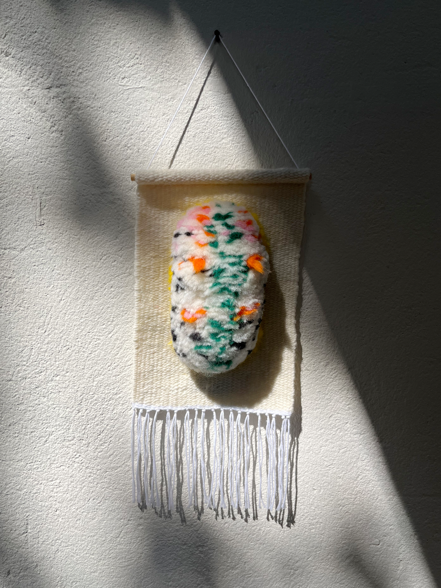 Little Crawler handwoven wall hanging with speckled sea slug creature in green, orange, and pink, hanging on a textured wall