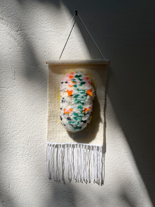 Little Crawler handwoven wall hanging with speckled sea slug creature in green, orange, and pink, hanging on a textured wall