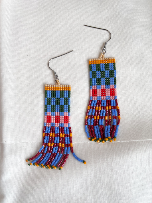 A pair of handmade beaded dangle earrings with a geometric pattern in blue, green, red, pink, and yellow, featuring silver-toned fishhook wires, displayed on a textured white fabric background.