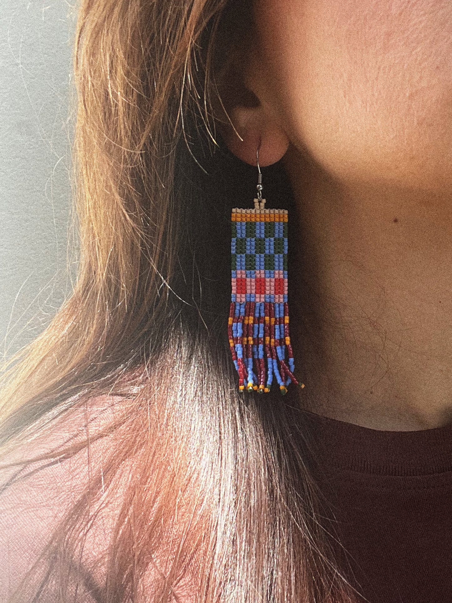 A close-up of a person wearing a long rectangular beaded dangle earring. The earring features geometric patterns in blue, red, orange, yellow, and white, hanging from a silver fishhook ear wire.