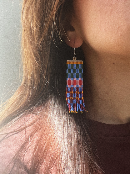 A close-up of a person wearing a long rectangular beaded dangle earring. The earring features geometric patterns in blue, red, orange, yellow, and white, hanging from a silver fishhook ear wire.