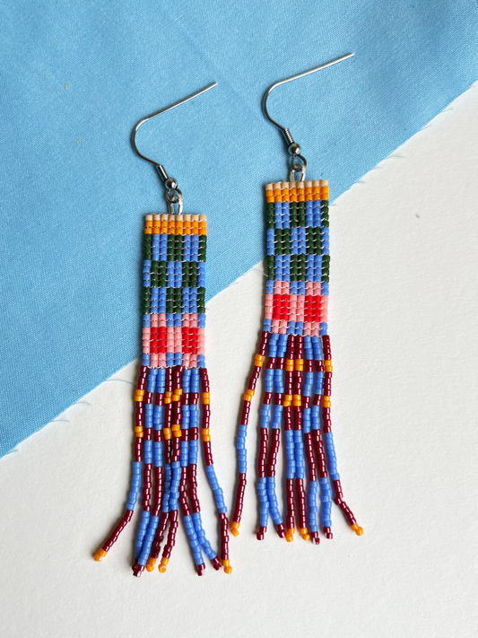 A pair of handmade beaded dangle earrings with fishhook backs, featuring geometric patterns in blue, green, red, pink, orange, and yellow with long fringe tassels, displayed on a diagonally split light blue and white background.