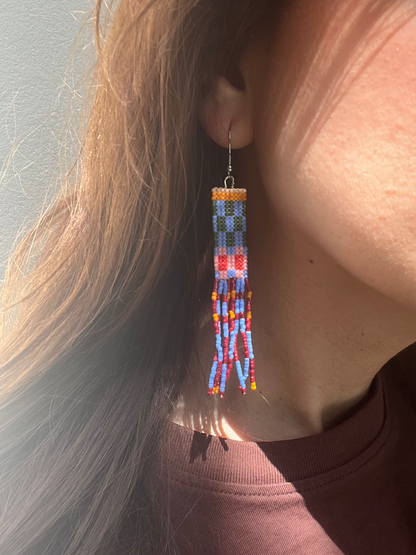 A close-up lifestyle shot of a person wearing a long, colorful beaded tassel earring. The earring features a geometric pattern in blue, red, orange, and yellow beads with dangling tassels.