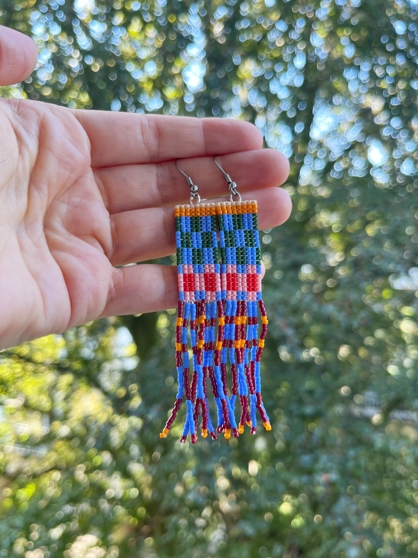A hand holds a pair of colorful beaded fringe earrings with silver fishhook wires against a blurred outdoor background of green trees.