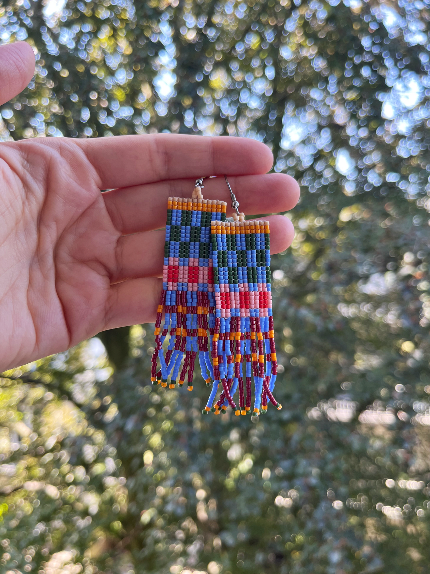 A pair of handmade rectangular beaded earrings held in a person's hand, featuring a colorful geometric pattern with blue, red, orange, yellow, and green beads, set against a blurred outdoor background of green trees.