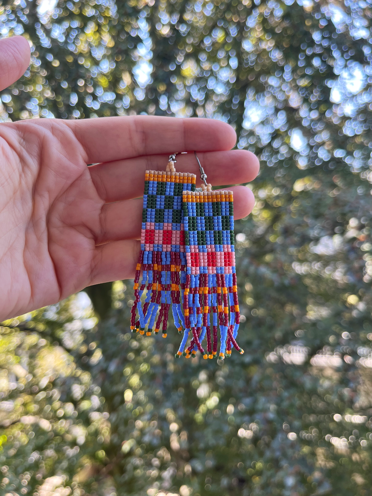 A pair of handmade rectangular beaded earrings held in a person's hand, featuring a colorful geometric pattern with blue, red, orange, yellow, and green beads, set against a blurred outdoor background of green trees.