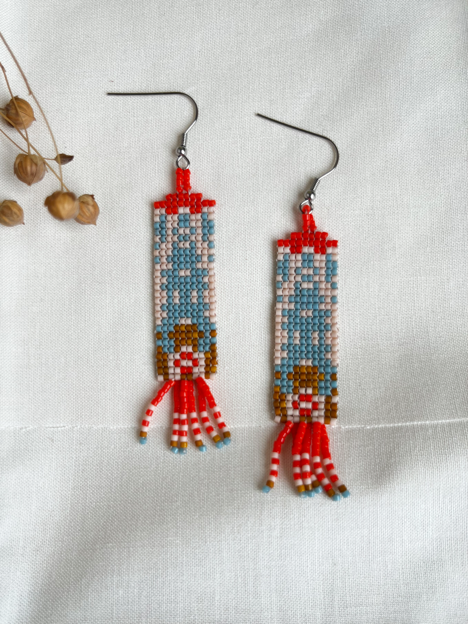 A pair of handmade beaded dangle earrings featuring a stylized design, displayed on a textured white fabric background with dried flower buds as props.
