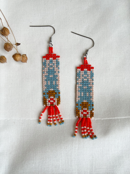 A pair of handmade beaded dangle earrings featuring a stylized design, displayed on a textured white fabric background with dried flower buds as props.