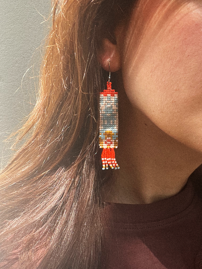 A close-up of a person wearing a long beaded dangle earring. The earring features a cylindrical pattern in red, white, and blue-gray beads, with a small gold-toned figure detail and red fringe at the bottom.