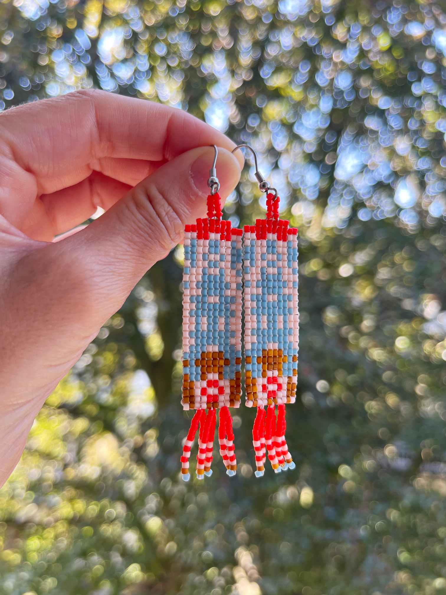 A hand holds a pair of long beaded dangle earrings outdoors against a blurred green and blue bokeh background. The earrings feature red, light blue, pink, and brown geometric patterns with red and white beaded fringe.