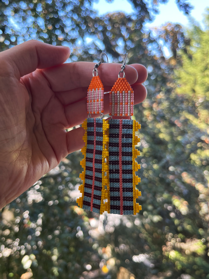 A hand holds a pair of long beaded dangle earrings outdoors with a blurred green forest background. The earrings feature orange and white beaded tops with long rectangular drops in black, gray, and yellow with red accent lines.
