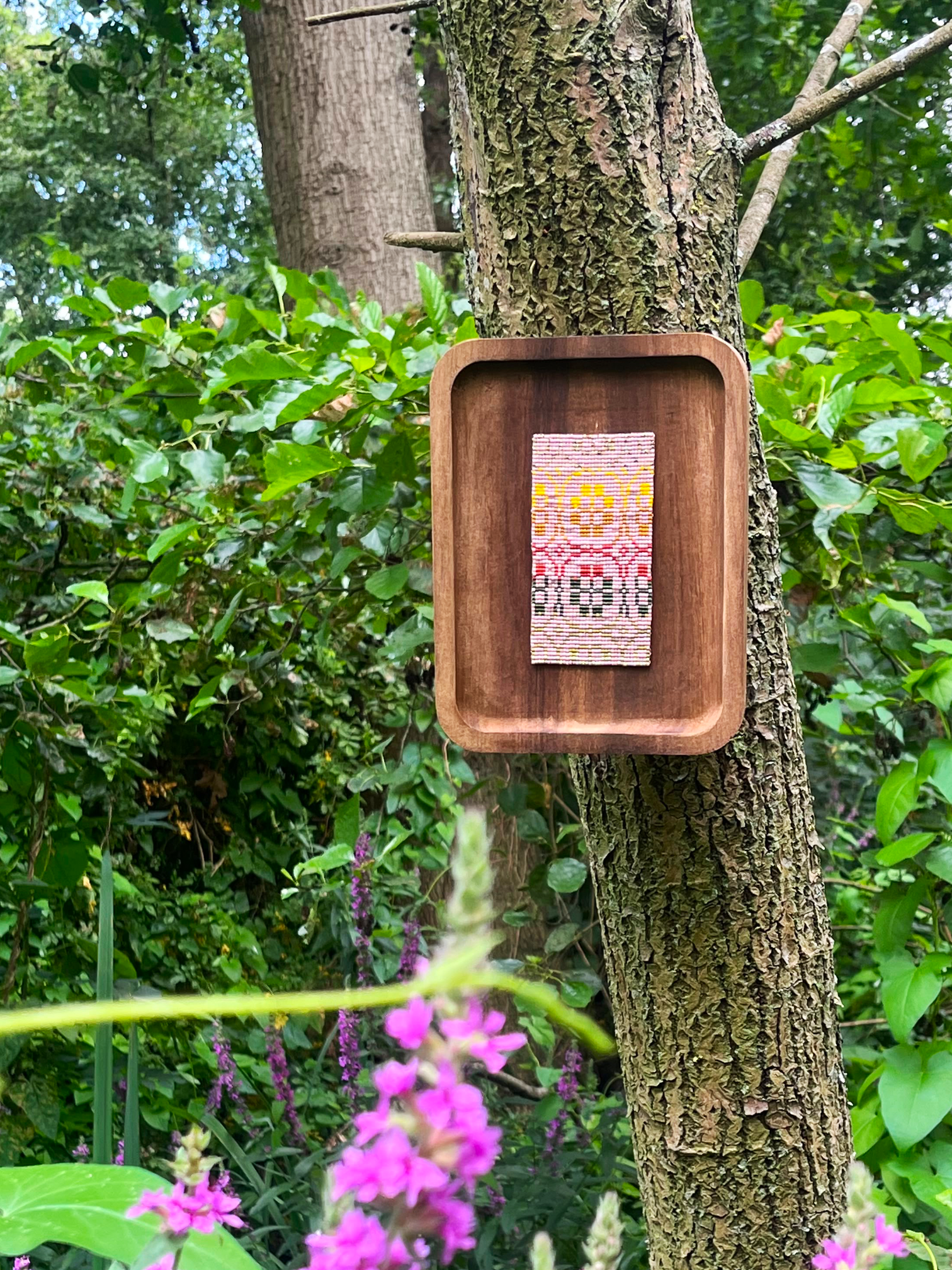 Handwoven beaded wall art hanging on a tree trunk in a garden with purple flowers