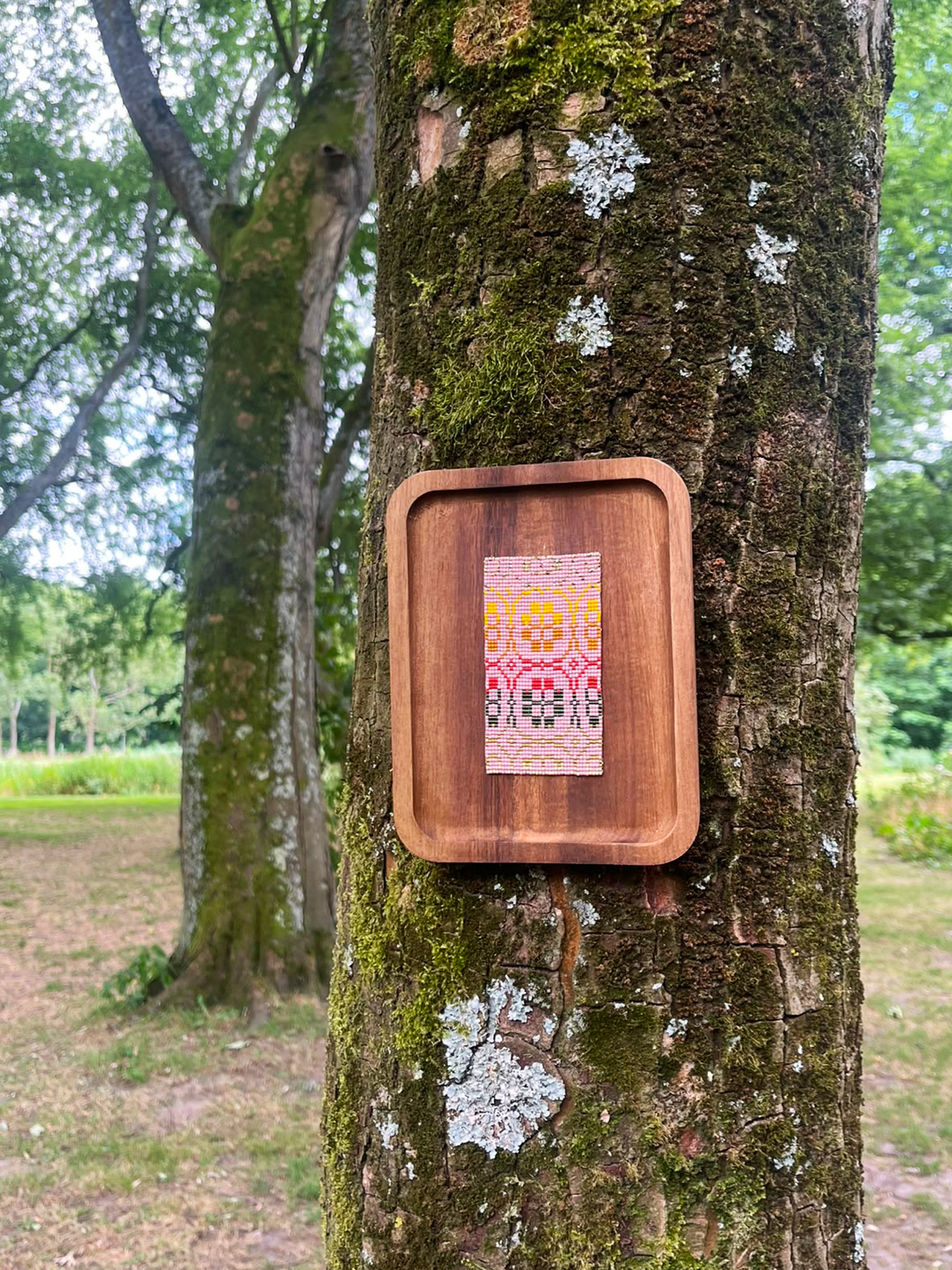 Floral beaded panel in wood frame hanging on a tree trunk in an open field setting