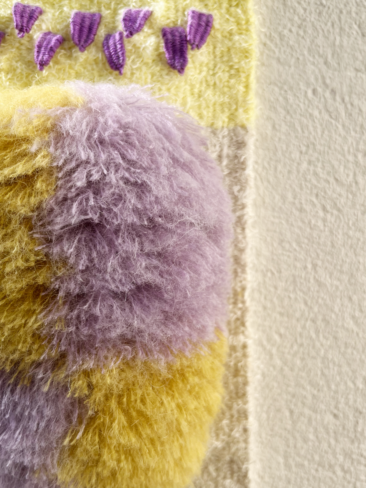 Close-up of a fuzzy, textured textile product in yellow and lavender-purple tones, with small purple stitched details near the top, shown against a plain off-white wall.
