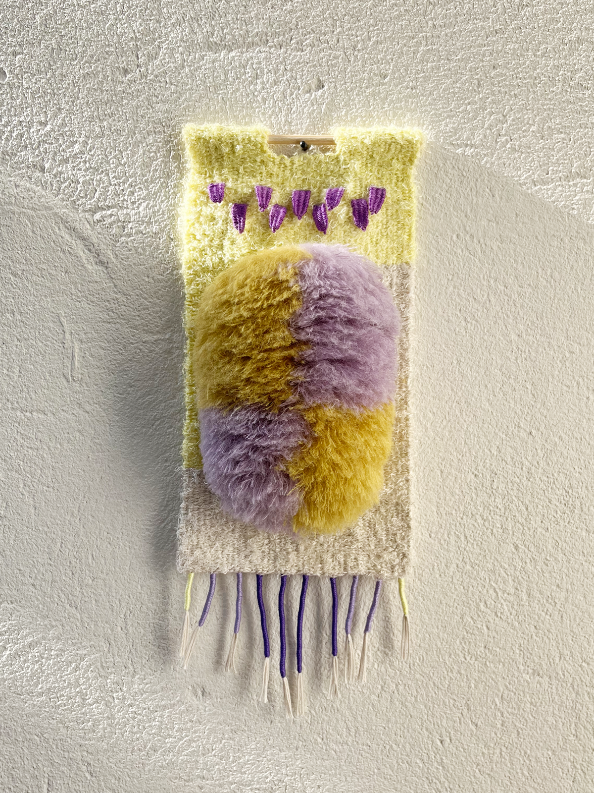 A handmade, textured wall hanging featuring a fuzzy, dual-toned oval in yellow and lavender, with purple tassels at the top and hanging fringes at the bottom, displayed against a textured off-white wall.