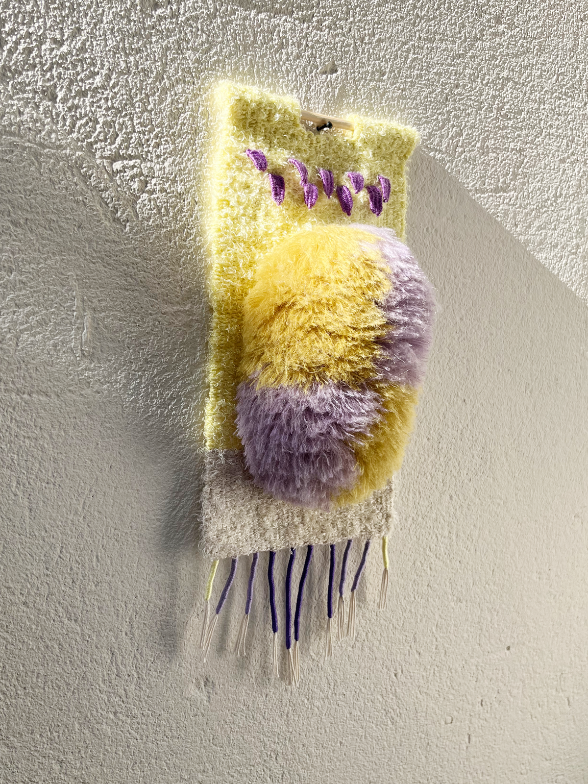 A handmade, textured wall hanging or decorative piece featuring fuzzy yellow and lavender purple loops, purple tassels, and white fringe, mounted on a textured off-white plaster wall.