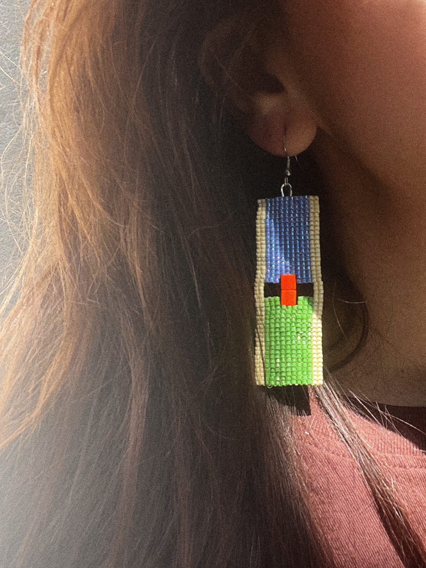 A close-up of a person wearing a long rectangular beaded dangle earring. The earring features a blue top section, a beige vertical border, a green lower section, and a small bright orange-red rectangular accent in the center.