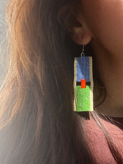 A close-up of a person wearing a long rectangular beaded dangle earring. The earring features a blue top section, a beige vertical border, a green lower section, and a small bright orange-red rectangular accent in the center.