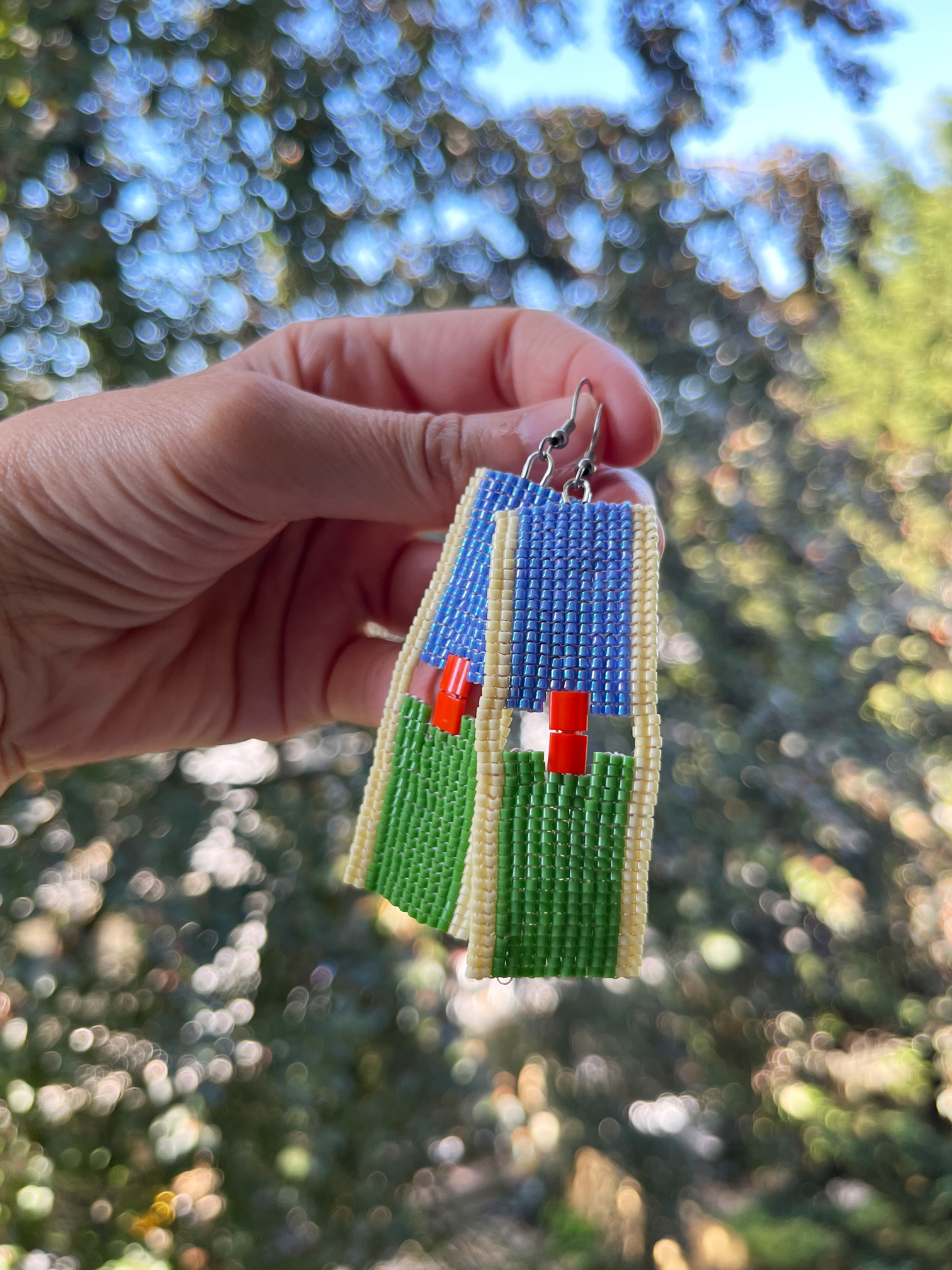 A hand holds a pair of handmade beaded earrings outdoors with a blurred forest background. The earrings feature a blue top section, a green bottom section, and small red and white bead accents, with cream-colored borders.
