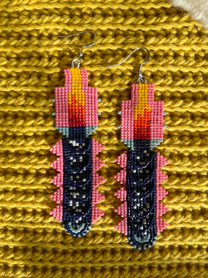 Dangling beaded earring. The earring is designed to look like a stylized candle, with a dark navy blue beaded body, pink beaded sides, a yellow and orange flame at the top, and a silver-tone hook.