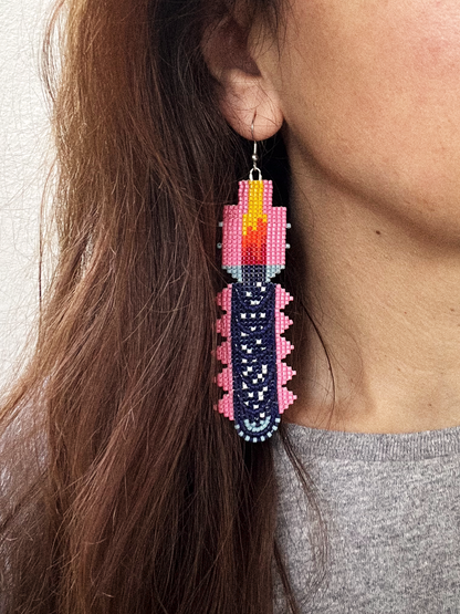 A close-up of a person wearing a long, dangling beaded earring. The earring is designed to look like a stylized candle, with a dark navy blue beaded body, pink beaded sides, a yellow and orange flame at the top, and a silver-tone hook.