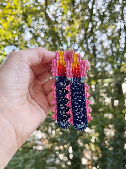 A hand holding a pair of colorful beaded earrings outdoors with a blurred green forest background. The earrings feature a geometric design in pink, navy blue, yellow, and red.