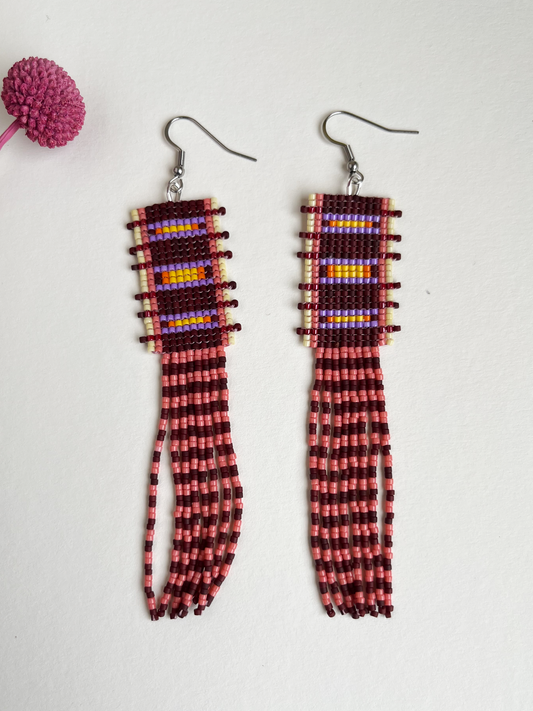 A pair of long beaded dangle earrings with silver fishhook backs, featuring a geometric patterned top section and long fringe below, displayed on a light off-white background next to a small pink decorative pom-pom.