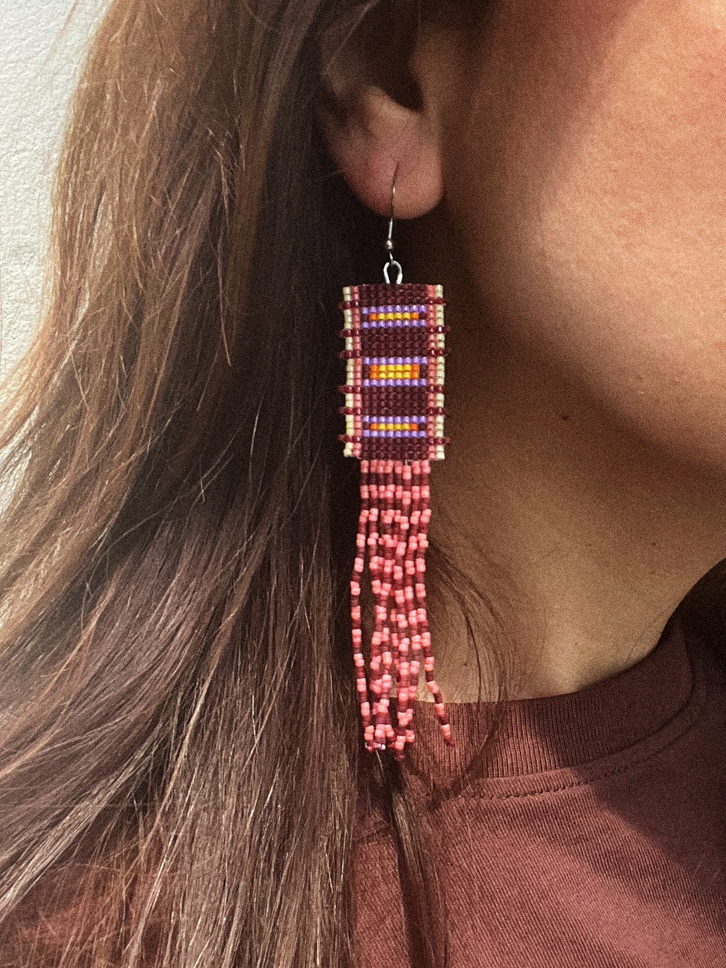 A close-up of a person wearing a long beaded dangle earring. The earring features a rectangular top section with purple, red, and yellow patterned beads, hanging from a silver fishhook wire, with multiple strands of red and pink beads dangling below.